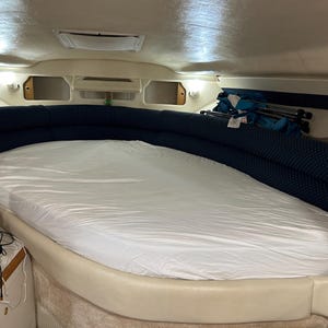 May include: Interior of a boat cabin featuring a large, white-sheeted bed. The bed is surrounded by a curved, navy blue upholstered headboard with a dotted pattern. Overhead lighting and a small side table with electronics are visible.