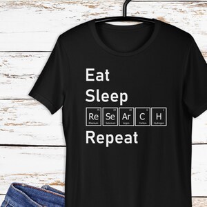Puede incluir: Camiseta negra con texto blanco que dice "Eat Sleep Research Repeat" con la palabra "Research" deletreada usando los símbolos químicos de Renio, Selenio, Argón, Carbono e Hidrógeno.