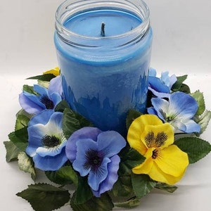 May include: A blue candle in a glass jar surrounded by a wreath of blue, purple, and yellow pansies.