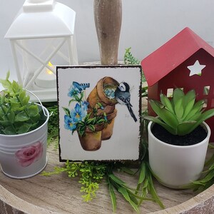 May include: A wooden tray with a white lantern, a red metal house, two potted plants, and a square tile with a bird and flowers. The tile has a blue bird with a nest in a pot, and blue and white flowers.
