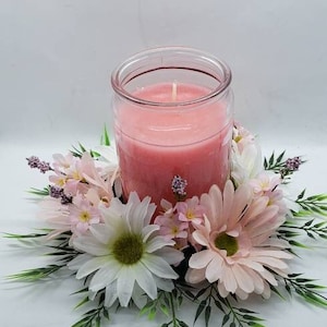 May include: A pink candle in a glass jar sits in the center of a floral arrangement. The arrangement features white and pink daisies, pink flowers, and green leaves.