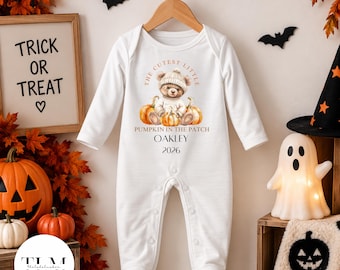 Puede incluir: Un pelele blanco de manga larga para bebé con pies, que presenta un gráfico de un osito de peluche con gorro sentado entre calabazas. El texto dice THE CUTEST LITTLE PUMPKIN IN THE PATCH, OAKLEY y 2026. La prenda tiene cierre de botones a presión.