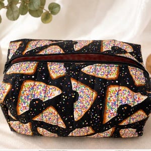 May include: A rectangular fabric pouch with a black background featuring a repeating pattern of triangular pastries topped with white icing and colourful sprinkles. The pouch has a black zip closure along the top edge and a quilted texture.