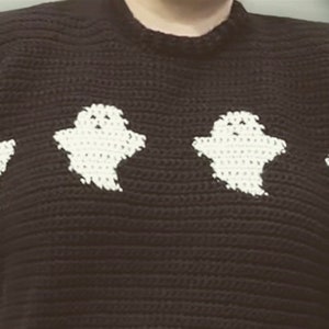 May include: Black crochet sweater featuring white ghost appliqués. The sweater has a crew neck and is made of a textured, dark yarn. The white ghosts are evenly spaced across the front of the sweater, creating a spooky, yet cute design.