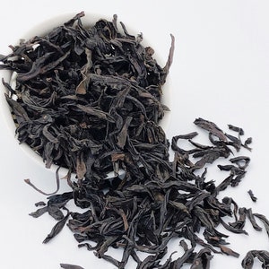 Könnte beinhalten: Lose schwarze Teeblätter in einer weißen Schüssel, die auf eine weiße Oberfläche fallen.