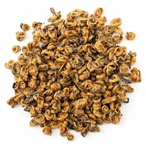 May include: A close-up shot of a pile of golden-brown tea leaves. The leaves are tightly curled and twisted, with a slightly darker hue in the center. The tea leaves are scattered on a white surface.