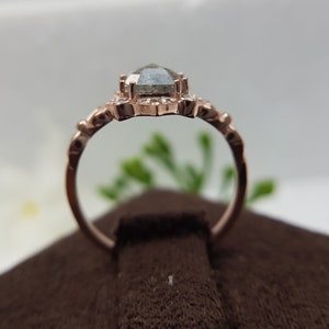 May include: A rose gold engagement ring with a gray, pear-shaped diamond set in a delicate floral halo. The ring is on a brown velvet surface.