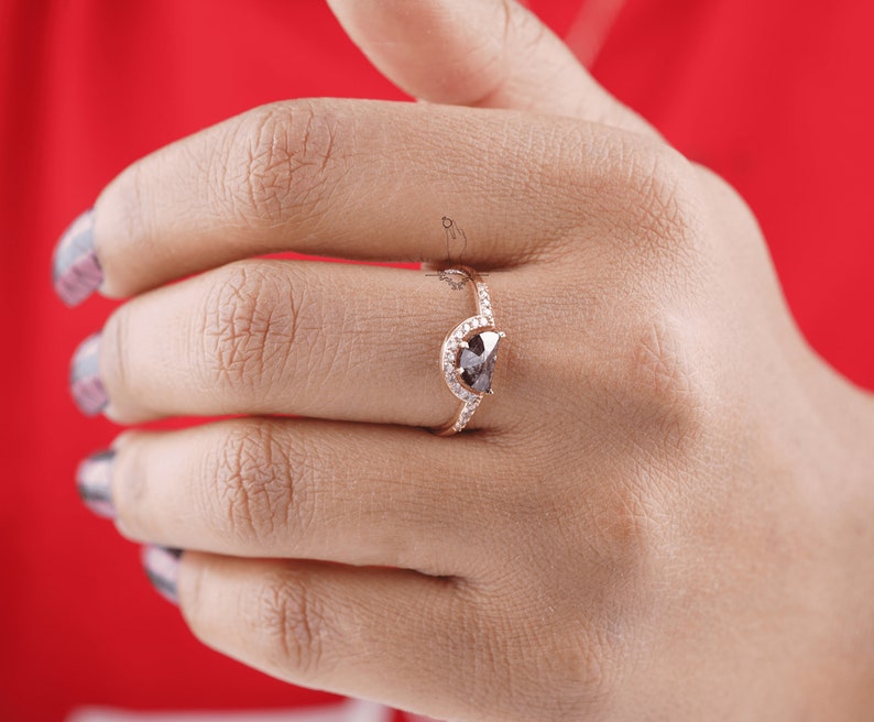 May include: A rose gold ring with a pear-shaped black diamond surrounded by smaller white diamonds. The ring is set on a half-circle band.
