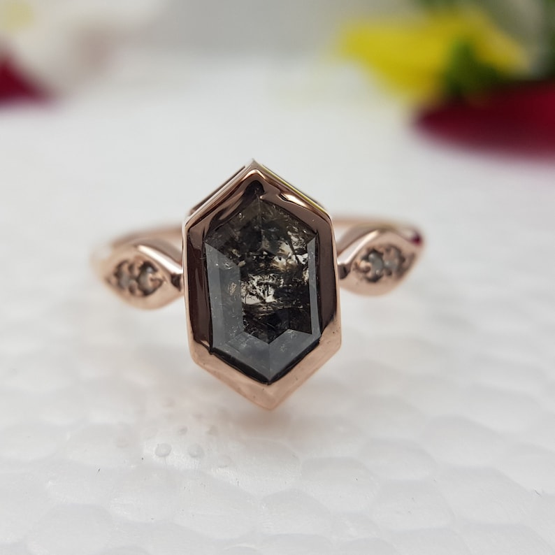 May include: A rose gold engagement ring with a hexagonal black diamond and two smaller diamonds on the band.