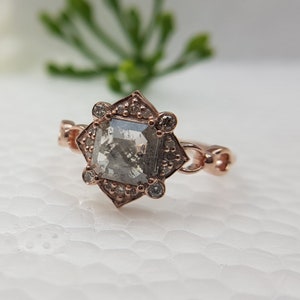 May include: A rose gold ring with a square-shaped, grey diamond set in a starburst-shaped halo. The ring has a delicate, chain-like band.