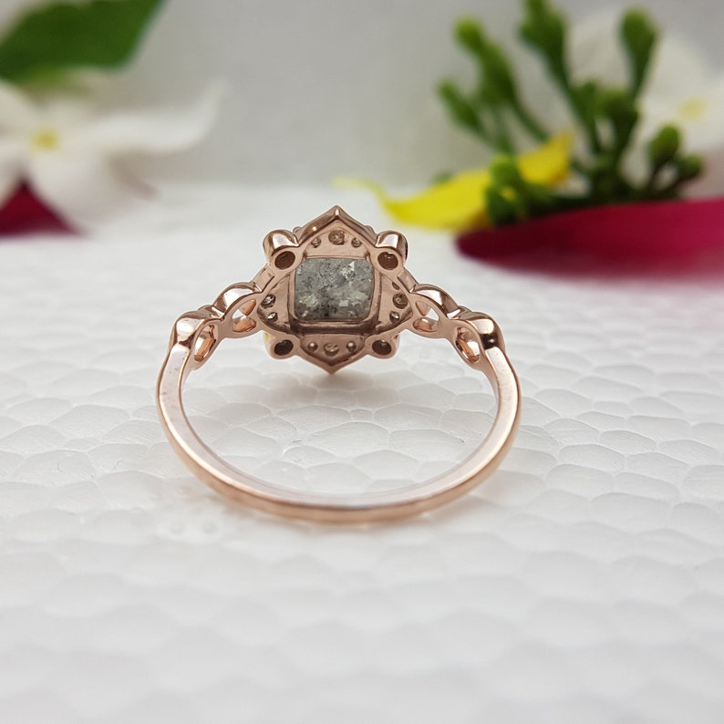 May include: A rose gold engagement ring with a square-shaped grey gemstone surrounded by smaller diamonds. The ring has a delicate, intricate design with a floral motif.