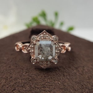 May include: A rose gold engagement ring with a square-shaped diamond surrounded by smaller diamonds. The ring has a delicate, intricate design.