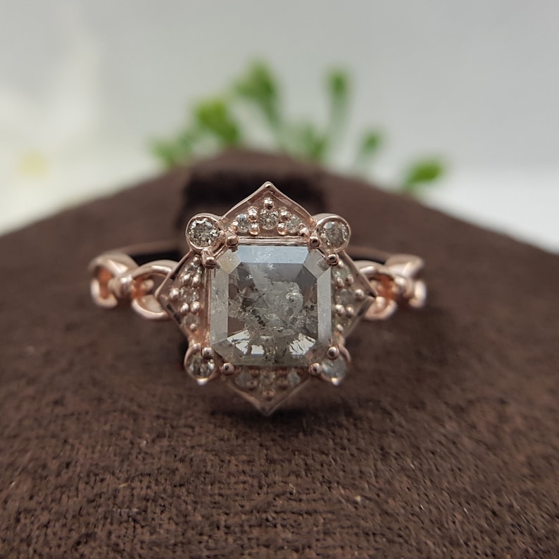 May include: A rose gold engagement ring with a square-shaped diamond surrounded by smaller diamonds. The ring has a delicate, intricate design.