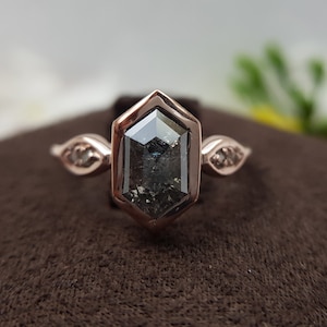 May include: A rose gold engagement ring with a hexagonal setting featuring a large, dark gray, salt and pepper diamond. The ring has two smaller diamonds on each side of the main stone.
