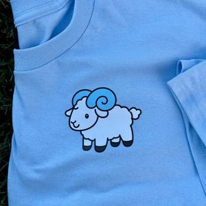 May include: Light blue t-shirt featuring a cartoon sheep graphic. The sheep is light blue with darker blue horns and black hooves. The shirt is made of a soft material and has a crew neck.