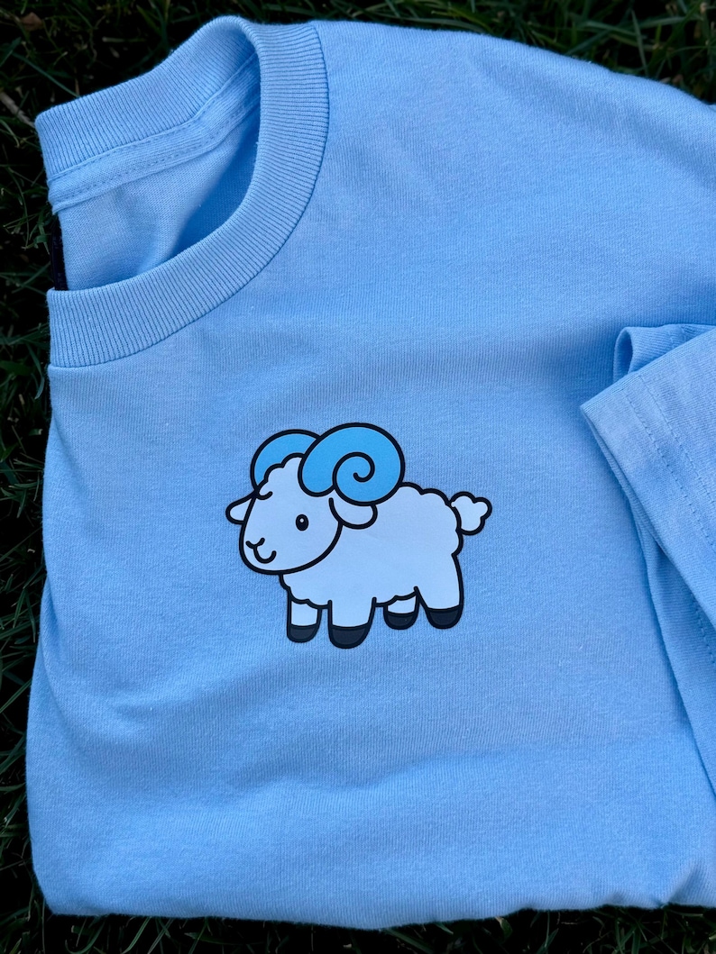 May include: Light blue t-shirt featuring a cartoon sheep graphic. The sheep is light blue with darker blue horns and black hooves. The shirt is made of a soft material and has a crew neck.