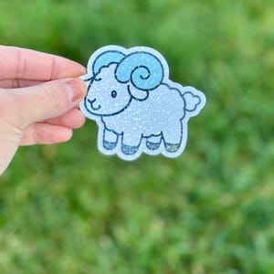 May include: A glittery sticker of a cartoon ram with blue horns and accents. The ram is white with glitter, and the sticker has a white border. The sticker is held against a blurred green background.