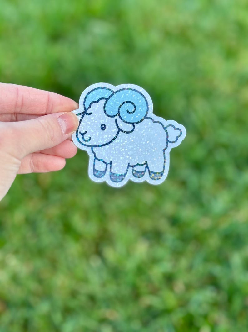 May include: A glittery sticker of a cartoon ram with blue horns and accents. The ram is white with glitter, and the sticker has a white border. The sticker is held against a blurred green background.