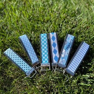 May include: Six blue key fobs with various patterns, including polka dots, argyle, and gingham, are arranged on green grass. Each fob has a silver-colored metal clasp and a dark blue fabric backing. One fob has the word "Carolina" printed on it.