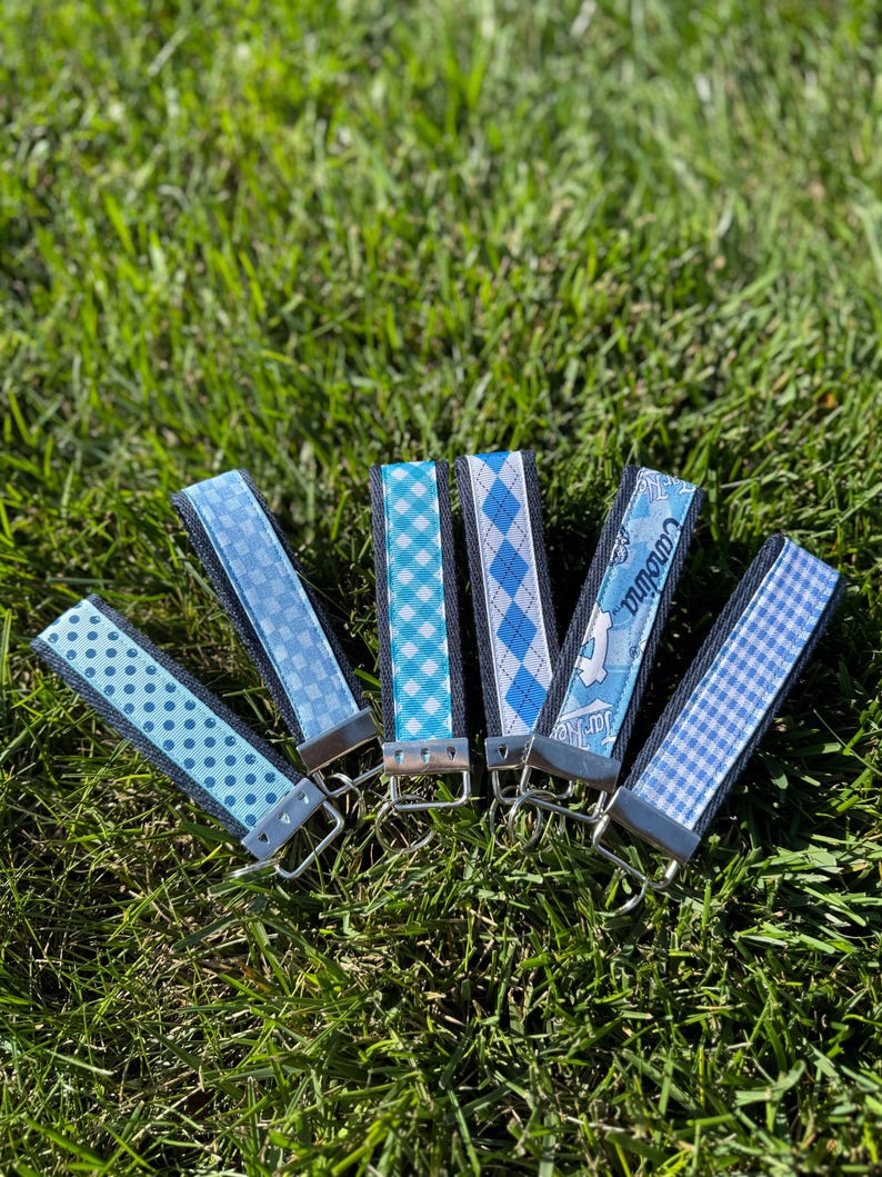 May include: Six blue key fobs with various patterns, including polka dots, argyle, and gingham, are arranged on green grass. Each fob has a silver-colored metal clasp and a dark blue fabric backing. One fob has the word "Carolina" printed on it.