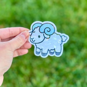 May include: A holographic sticker of a cartoon ram with blue horns and accents. The ram has a smiling face and is outlined in black. The sticker is held against a blurred green background.
