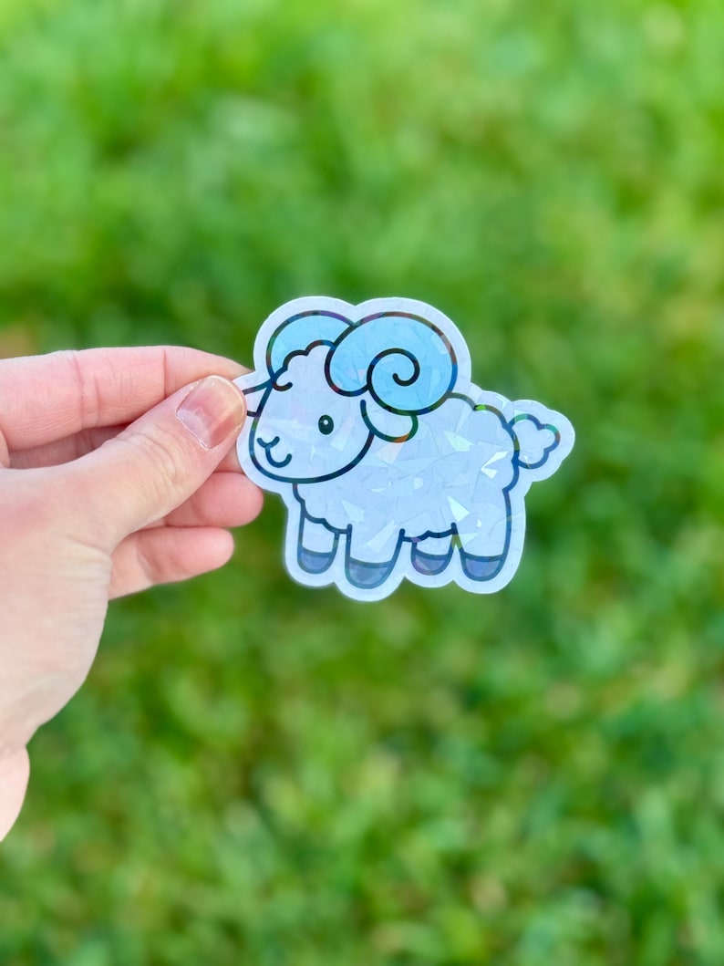 May include: A holographic sticker of a cartoon ram with blue horns and accents. The ram has a smiling face and is outlined in black. The sticker is held against a blurred green background.