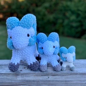 May include: Three handmade crochet plush toys in the shape of a ram. The toys are white with blue accents and gray hooves. The toys are arranged from largest to smallest, sitting on a wooden surface.