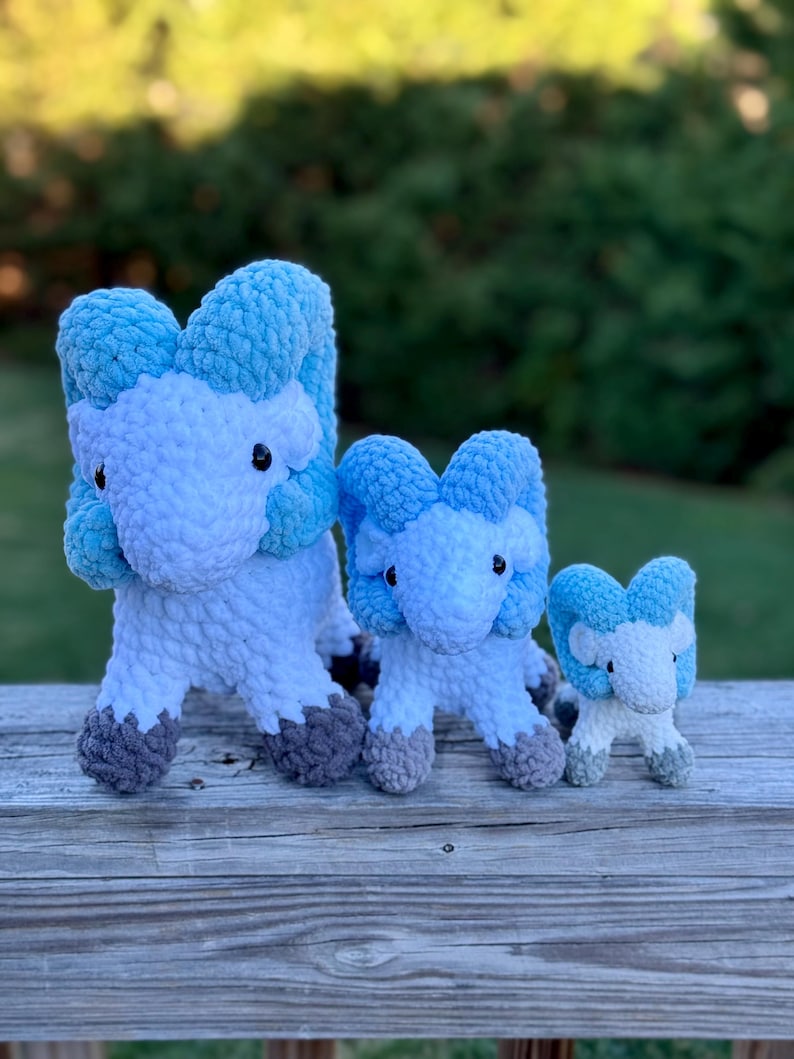 May include: Three handmade crochet plush toys in the shape of a ram. The toys are white with blue accents and gray hooves. The toys are arranged from largest to smallest, sitting on a wooden surface.