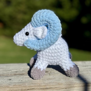 May include: A handmade crochet ram figurine. The ram is primarily white with light blue horns and accents. The hooves are gray. The ram is sitting on a wooden surface with a blurred green background.
