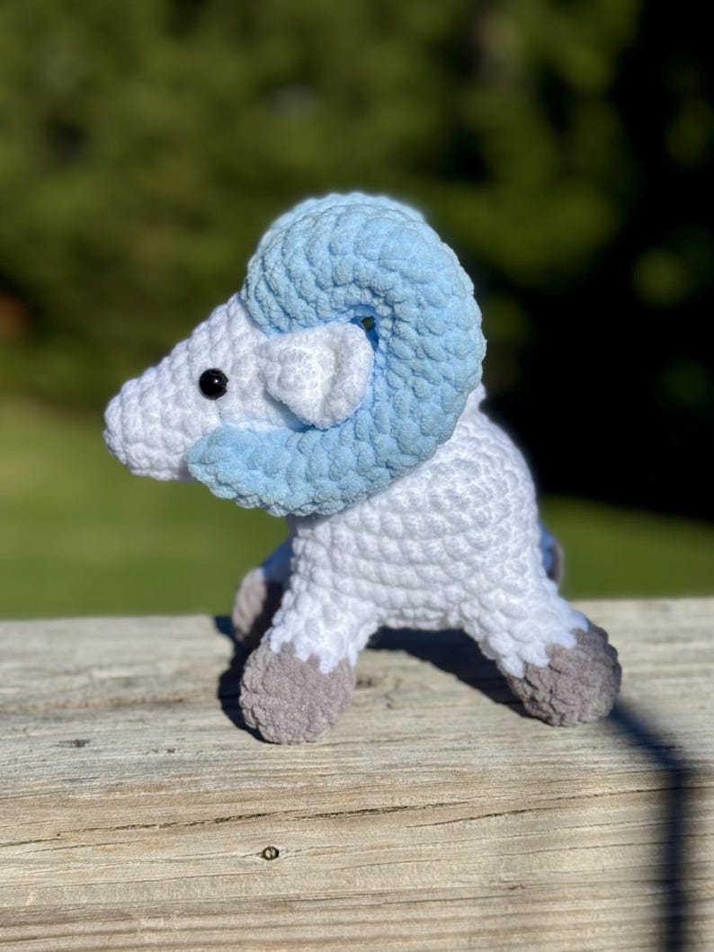May include: A handmade crochet ram figurine. The ram is primarily white with light blue horns and accents. The hooves are gray. The ram is sitting on a wooden surface with a blurred green background.