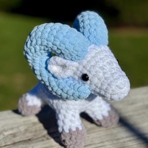 May include: A handmade crochet ram toy in shades of white, light blue, and gray. The ram has large, curved horns in light blue, a white body, and gray hooves. The toy is sitting on a wooden surface with a blurred green background.