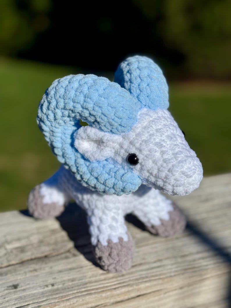 May include: A handmade crochet ram toy in shades of white, light blue, and gray. The ram has large, curved horns in light blue, a white body, and gray hooves. The toy is sitting on a wooden surface with a blurred green background.
