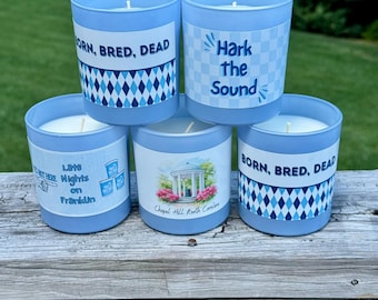May include: Five light blue candles with white wax and various designs. Some feature text like "BORN, BRED, DEAD" and "Hark the Sound." Others display diamond patterns and a Chapel Hill, North Carolina image. The candles are on a wooden surface.