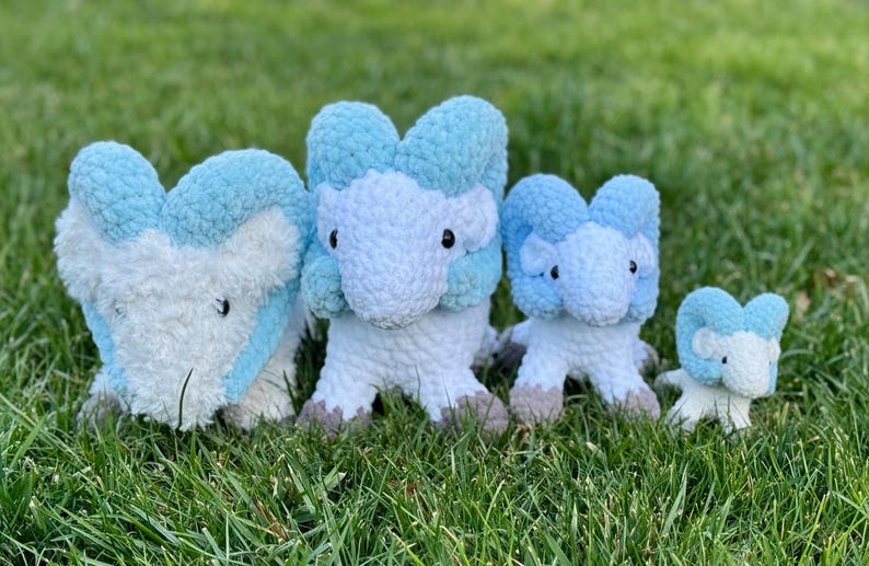 May include: Four plush toys in varying sizes, arranged on green grass. The toys are white with light blue accents and feature a stylized animal design. The largest toy is approximately 12 inches tall, with the smallest being about 4 inches.