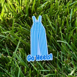 Puede incluir: Una pegatina con un dise&ntilde;o de peto a rayas azules y blancas con el texto "Go Heels!" en azul. La pegatina est&aacute; colocada sobre hierba verde.