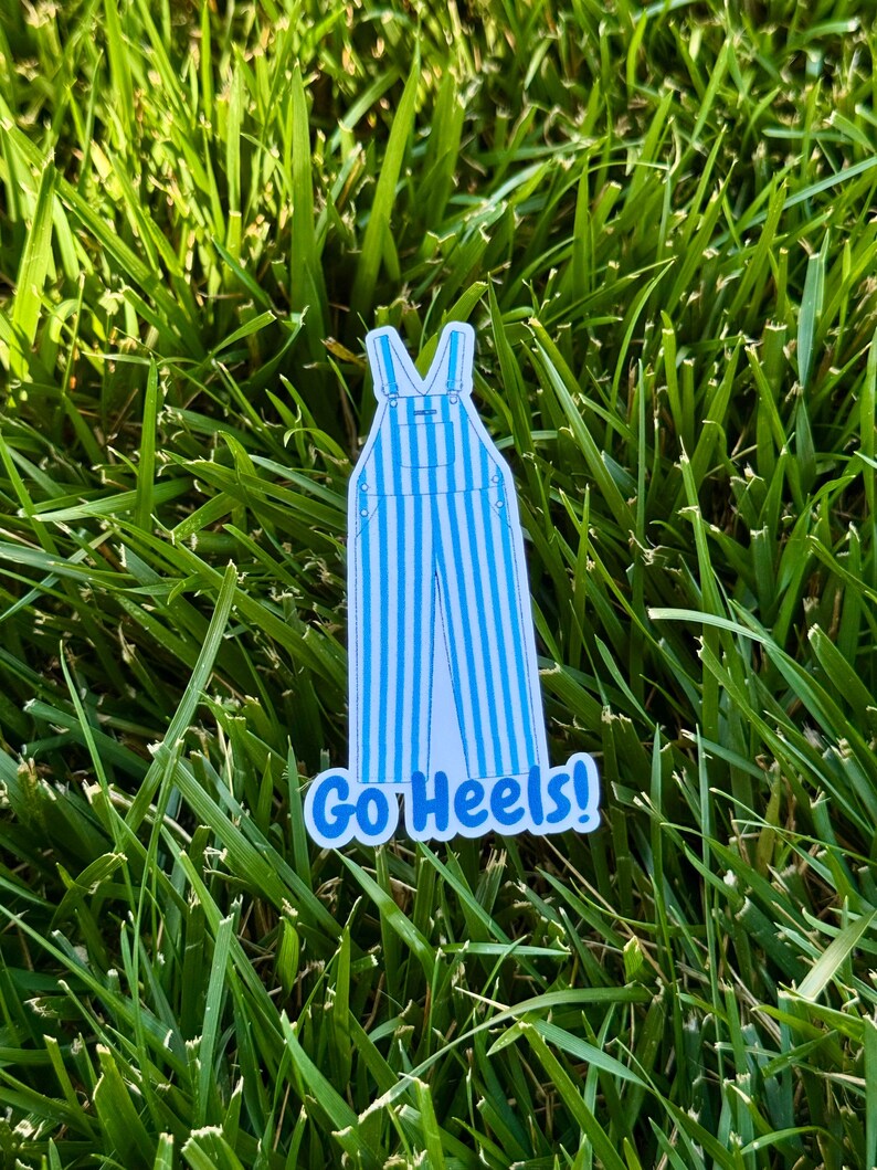 Puede incluir: Una pegatina con un dise&ntilde;o de peto a rayas azules y blancas con el texto "Go Heels!" en azul. La pegatina est&aacute; colocada sobre hierba verde.