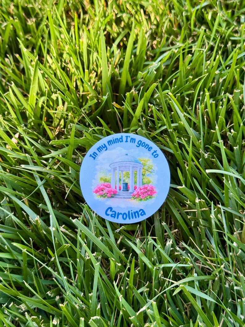 Puede incluir: Una pegatina redonda de color azul claro con el texto "In my mind I'm gone to Carolina". La pegatina presenta una ilustraci&oacute;n de un mirador con flores rosas y la palabra "Carolina" debajo. La pegatina est&aacute; sobre hierba verde.