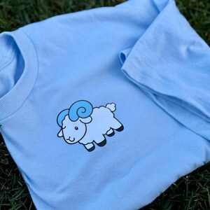 May include: A light blue long-sleeve shirt featuring a cartoon sheep graphic. The sheep is light blue with darker blue horns and black hooves. The shirt is made of a soft material and is suitable for casual wear.