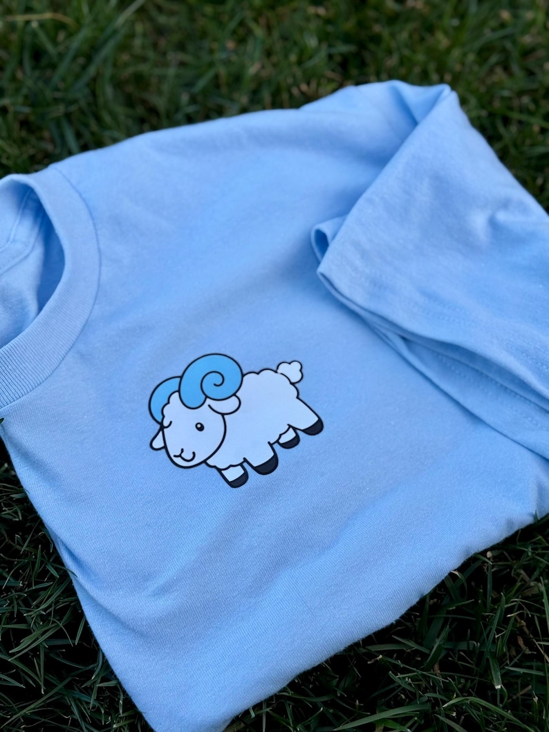 May include: A light blue long-sleeve shirt featuring a cartoon sheep graphic. The sheep is light blue with darker blue horns and black hooves. The shirt is made of a soft material and is suitable for casual wear.