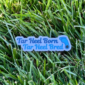 Puede incluir: Una pegatina rectangular con el texto "Tar Heel Born Tar Heel Bred" en azul claro, con un gr&aacute;fico de huella de beb&eacute;. La pegatina est&aacute; sobre un fondo de hierba verde.