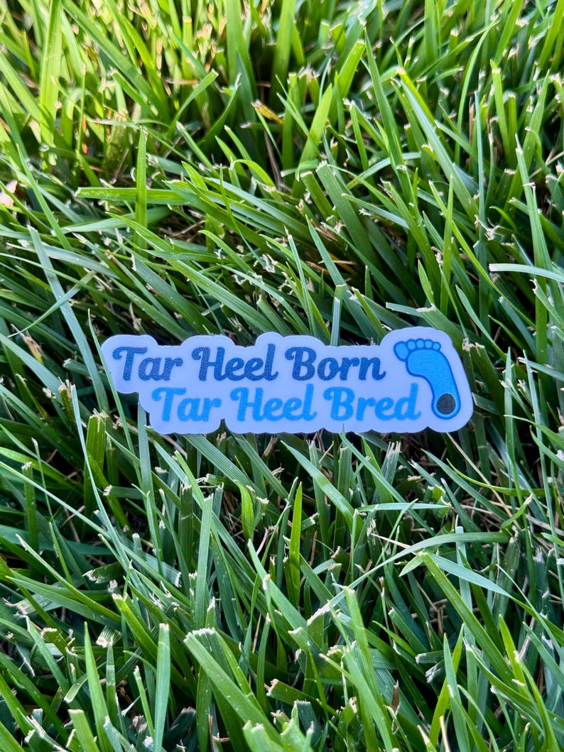 Puede incluir: Una pegatina rectangular con el texto "Tar Heel Born Tar Heel Bred" en azul claro, con un gr&aacute;fico de huella de beb&eacute;. La pegatina est&aacute; sobre un fondo de hierba verde.