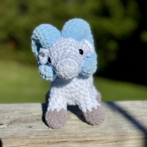 May include: A handmade crochet elephant toy in shades of blue, white, and gray. The elephant has large, floppy ears and small black eyes. The feet are a contrasting gray color. The toy is sitting on a wooden surface.