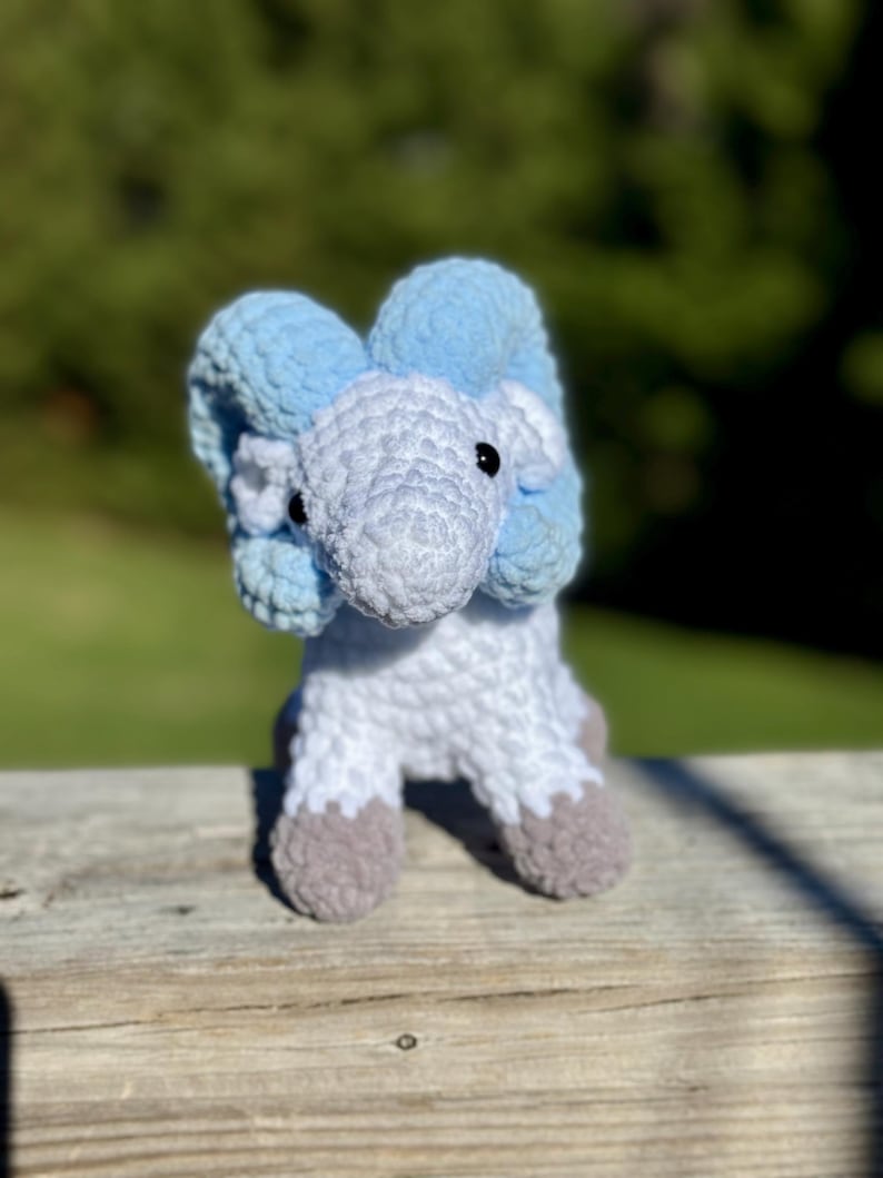 May include: A handmade crochet elephant toy in shades of blue, white, and gray. The elephant has large, floppy ears and small black eyes. The feet are a contrasting gray color. The toy is sitting on a wooden surface.