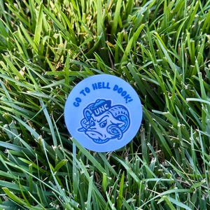 Puede incluir: Una pegatina redonda azul claro con el texto blanco "GO TO HELL DOOK!" que rodea una ilustraci&oacute;n azul y blanca de una cabeza de carnero con "UNC". La pegatina est&aacute; sobre hierba verde.