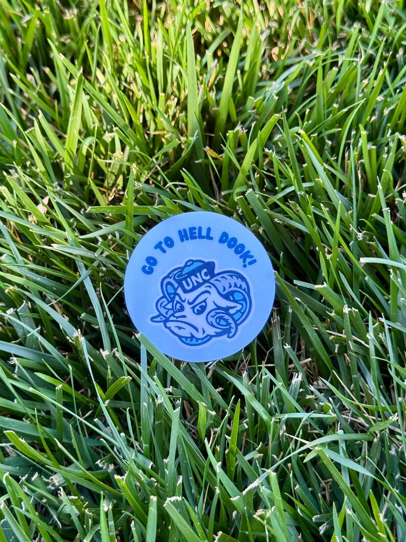 Puede incluir: Una pegatina redonda azul claro con el texto blanco "GO TO HELL DOOK!" que rodea una ilustraci&oacute;n azul y blanca de una cabeza de carnero con "UNC". La pegatina est&aacute; sobre hierba verde.