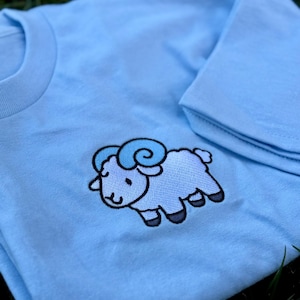 May include: Light blue t-shirt featuring an embroidered cartoon ram with light and dark blue accents. The ram has a smiling face and is outlined in black. The shirt is folded, with the collar and sleeve visible.