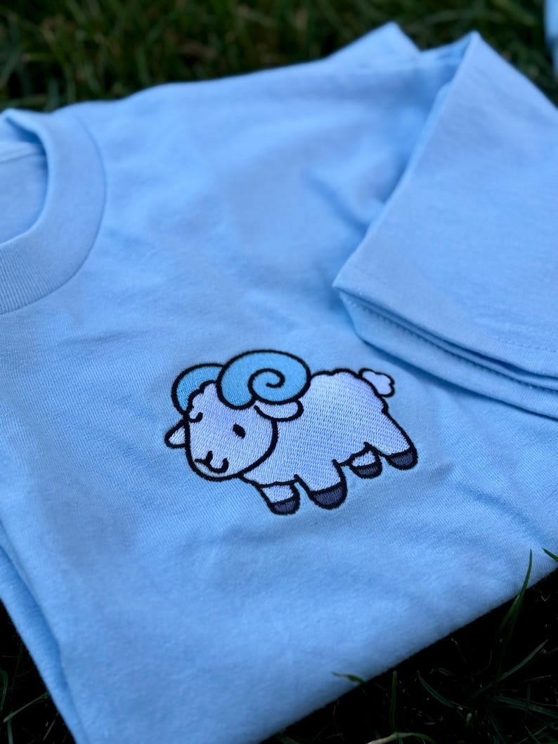 May include: Light blue t-shirt featuring an embroidered cartoon ram with light and dark blue accents. The ram has a smiling face and is outlined in black. The shirt is folded, with the collar and sleeve visible.
