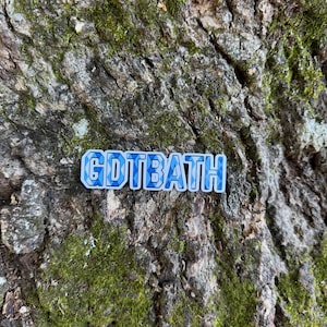 Puede incluir: Un pin rectangular con el texto "GDTBATH" en azul y blanco, sobre un fondo de corteza de &aacute;rbol texturizada y cubierta de musgo. Las letras del pin tienen un patr&oacute;n azul en forma de mosaico.