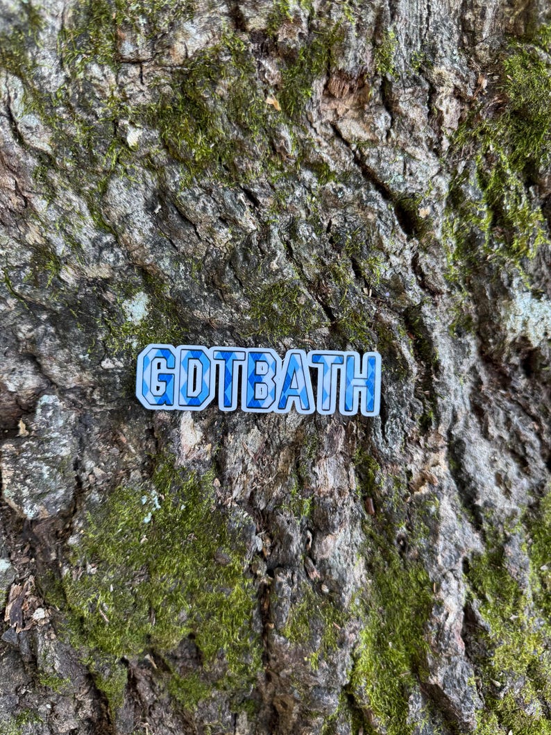 Puede incluir: Un pin rectangular con el texto "GDTBATH" en azul y blanco, sobre un fondo de corteza de &aacute;rbol texturizada y cubierta de musgo. Las letras del pin tienen un patr&oacute;n azul en forma de mosaico.
