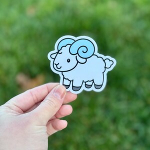 May include: A sticker depicting a cartoon ram with light blue fur and curly horns. The ram has black hooves and a smiling face. The sticker is on a white background.